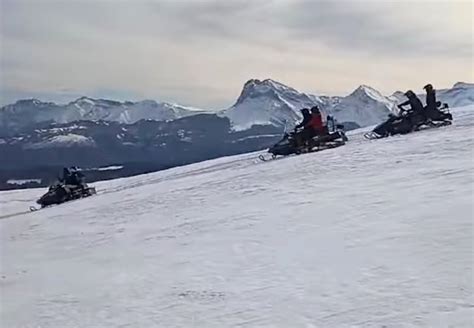 Panoramica di un gruppo di motoslitte che sfrecciano su un sentiero innevato in montagna, con alberi coperti di neve sullo sfondo.