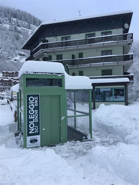Panorama alpino con l'Hotel Club Funivia in primo piano, circondato da prati verdi e montagne innevate.