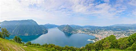 Vista panoramica di un lago alpino circondato da montagne verdi e cielo azzurro.