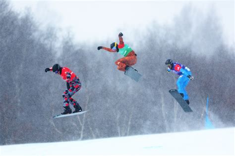 Gruppo di snowboarder che competono fianco a fianco in una gara di snowboard cross, con ostacoli sul percorso.